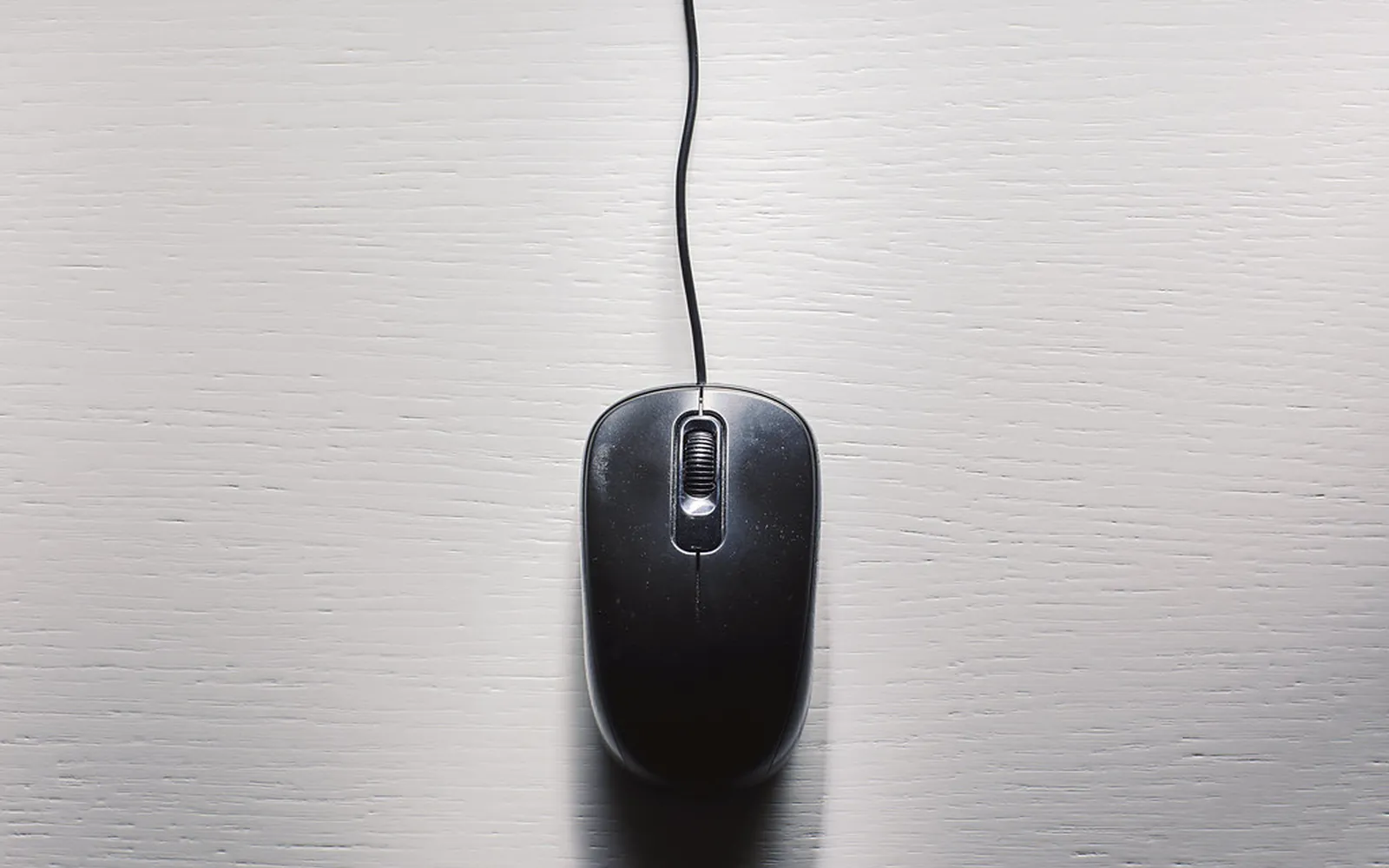 A black wired computer mouse on a pale wooden desk, seen from above